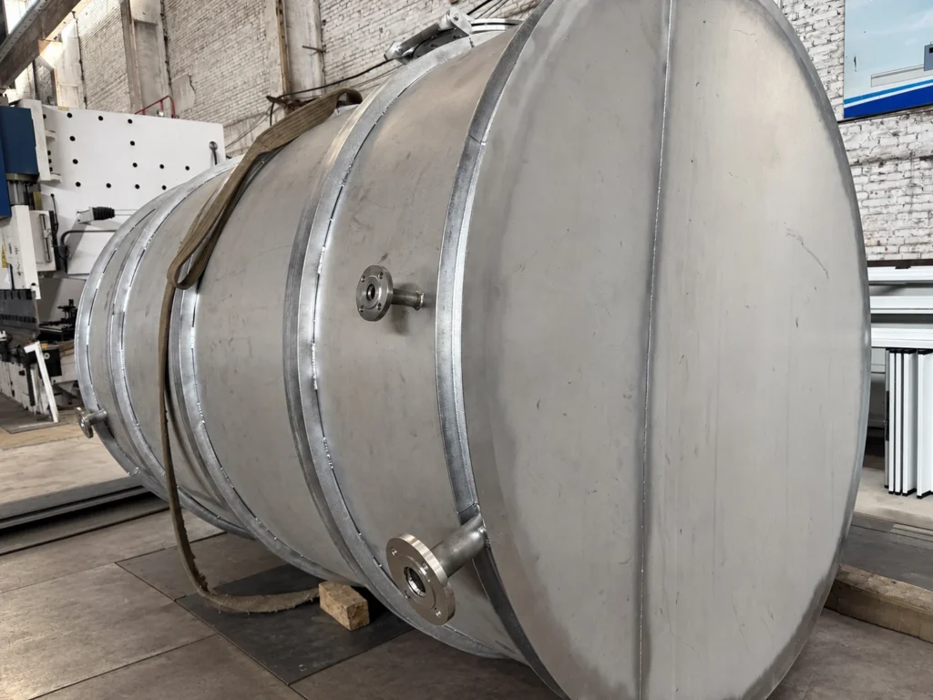 Fabricated stainless steel tank resting horizontally in the workshop, with nozzles and external rings